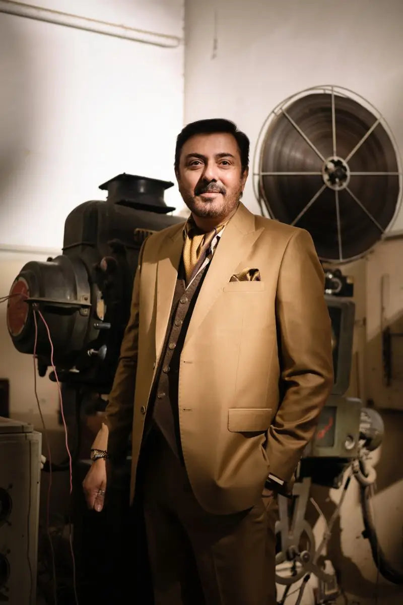 A man in a tan designer 3-piece suit for men stands confidently beside vintage film equipment in agha talish luxury pakistani groom wear