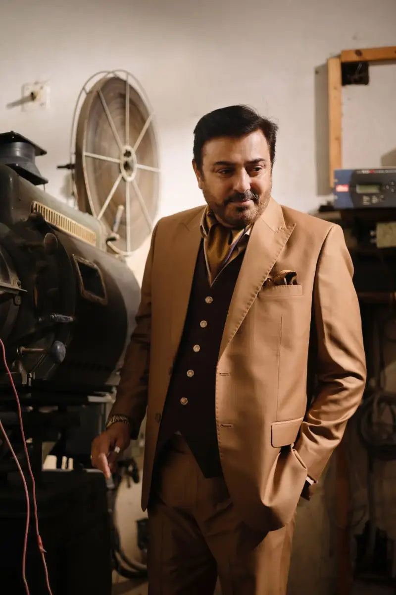 A man in a tan designer 3-piece suit for men stands beside vintage film equipment showcasing luxury pakistani groom wear
