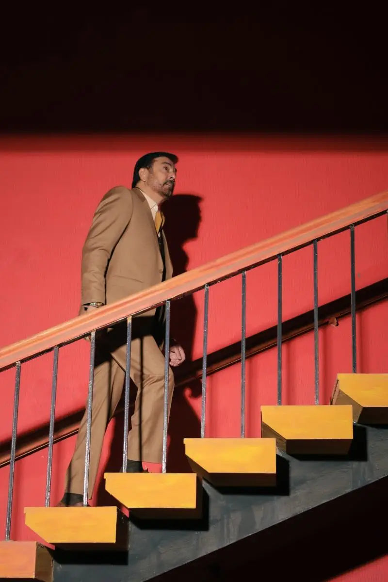 A man wearing a designer 3-piece tan suit with a yellow tie stands on bright orange stairs for luxury pakistani groom wear