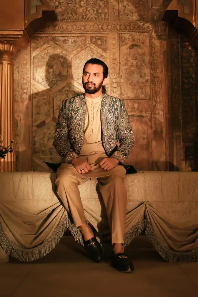 Man wearing beige paisley embroidered ghazal jacket and matching trousers seated on ornate draped bench