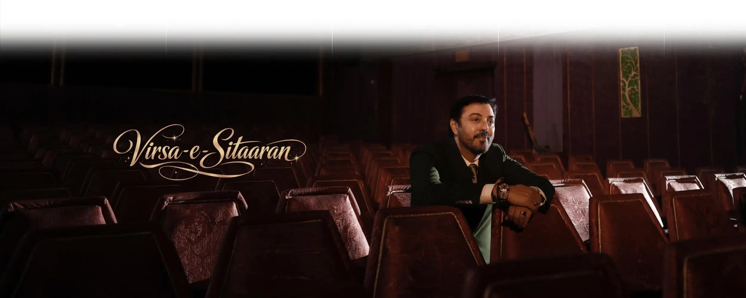 A man in a dark suit sits alone in an empty, dimly lit theater with rows of red upholstered seats.
