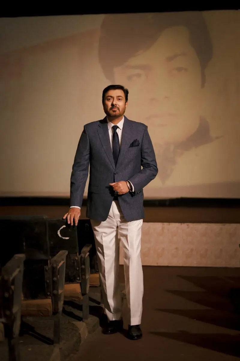 A man in a tailored navy blue suit with white trousers and black shoes standing by an empty theater seat for muhammad ali virsa-e-sitaaran edit26