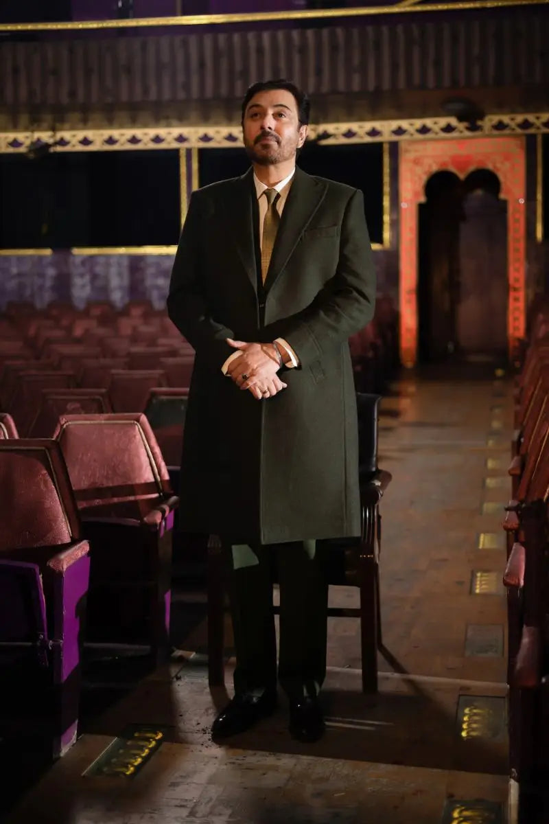 A man in a dark green overcoat with matching suit and tie stands clasping hands for nadeem baig virsa e sitaaran edit26