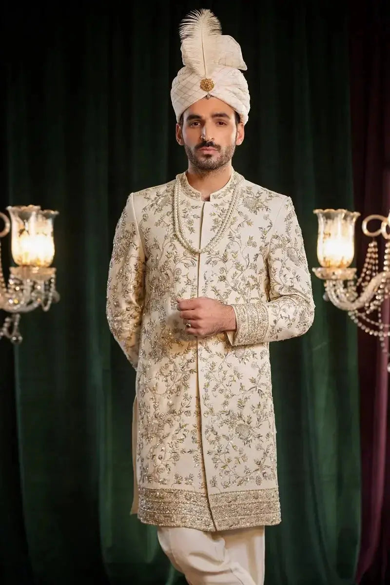 Cream-colored embroidered sherwani saleem with gold thread detailing and a matching turban adorned with a white feather