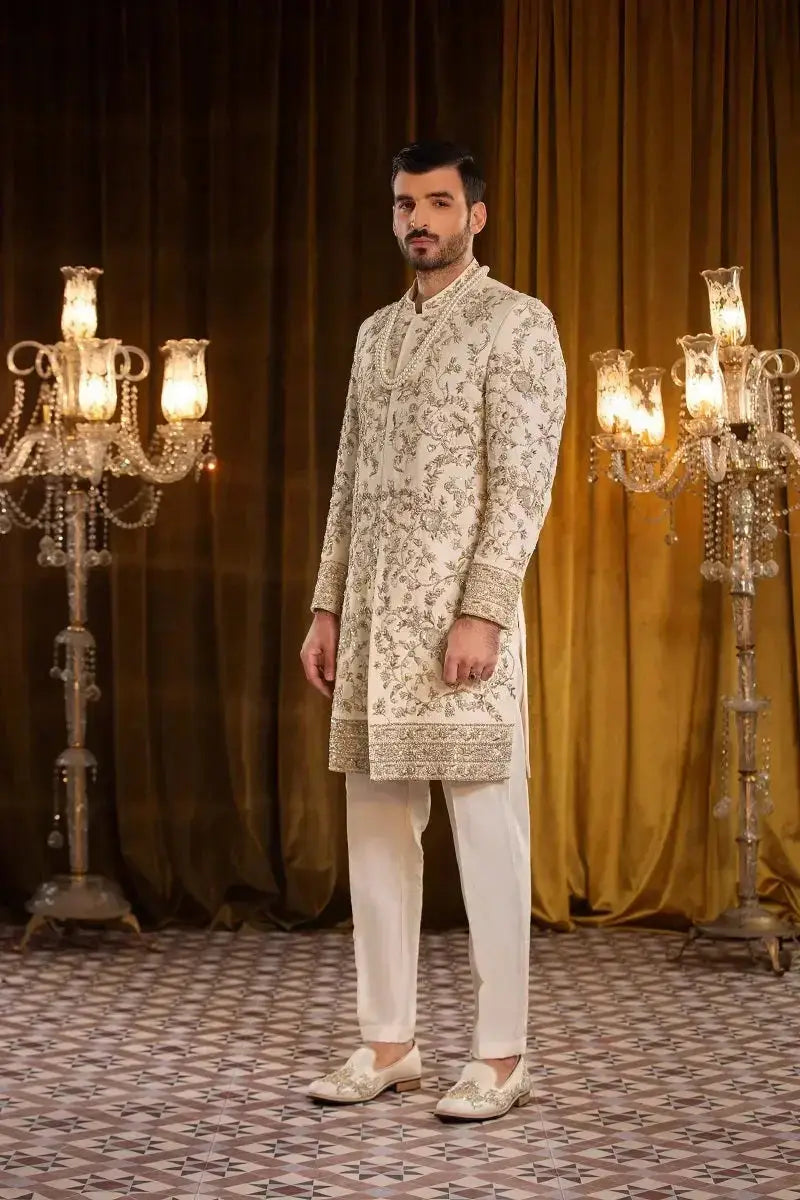 A man wearing a cream-colored sherwani with gold embroidery and white trousers in the SALEEM collection