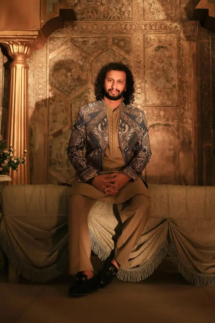 A man wearing a shayar jacket with shimmering silver and blue embroidery over beige trousers
