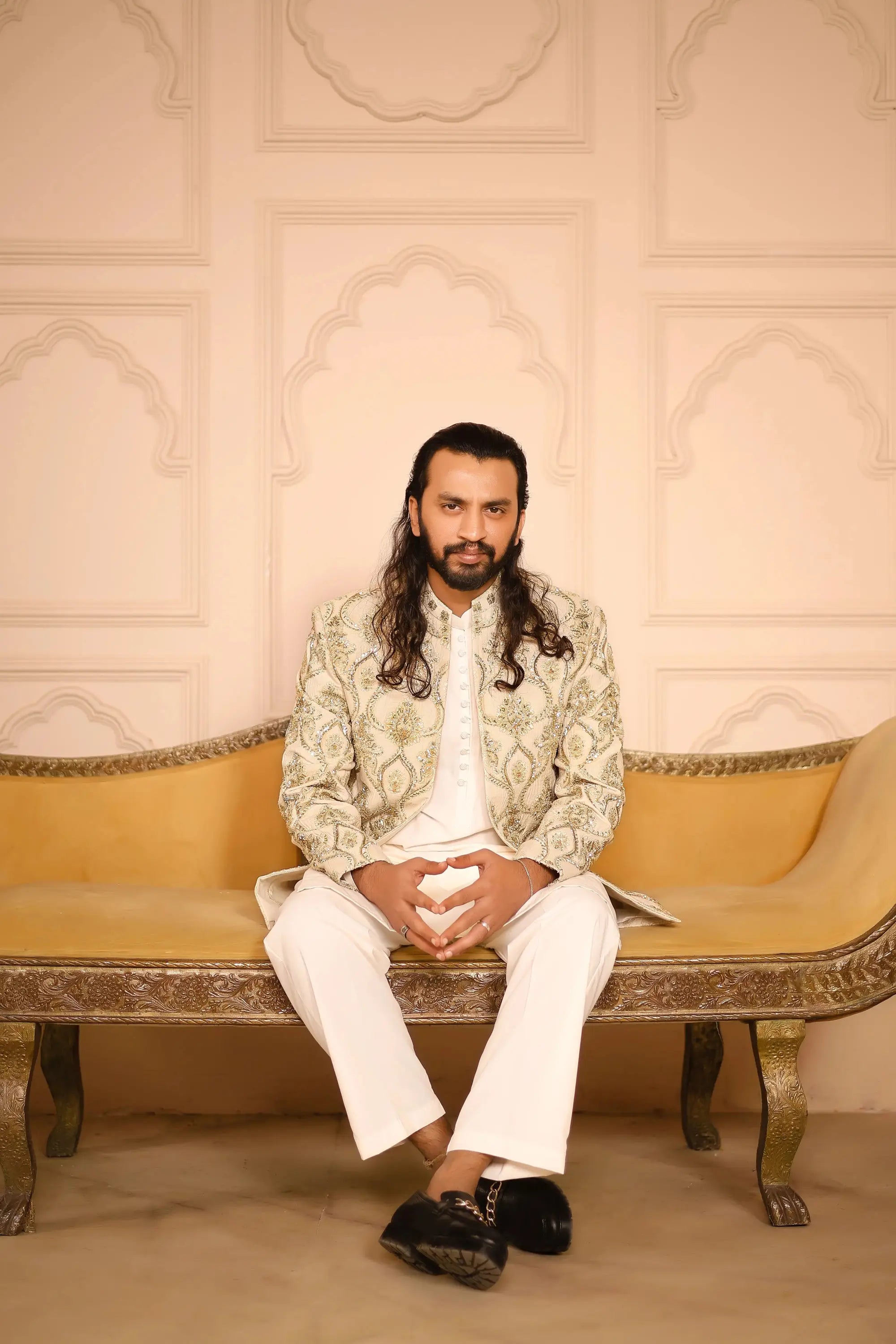 A man sits on a golden bench wearing a cream Sikandar jacket with green embroidery and white trousers