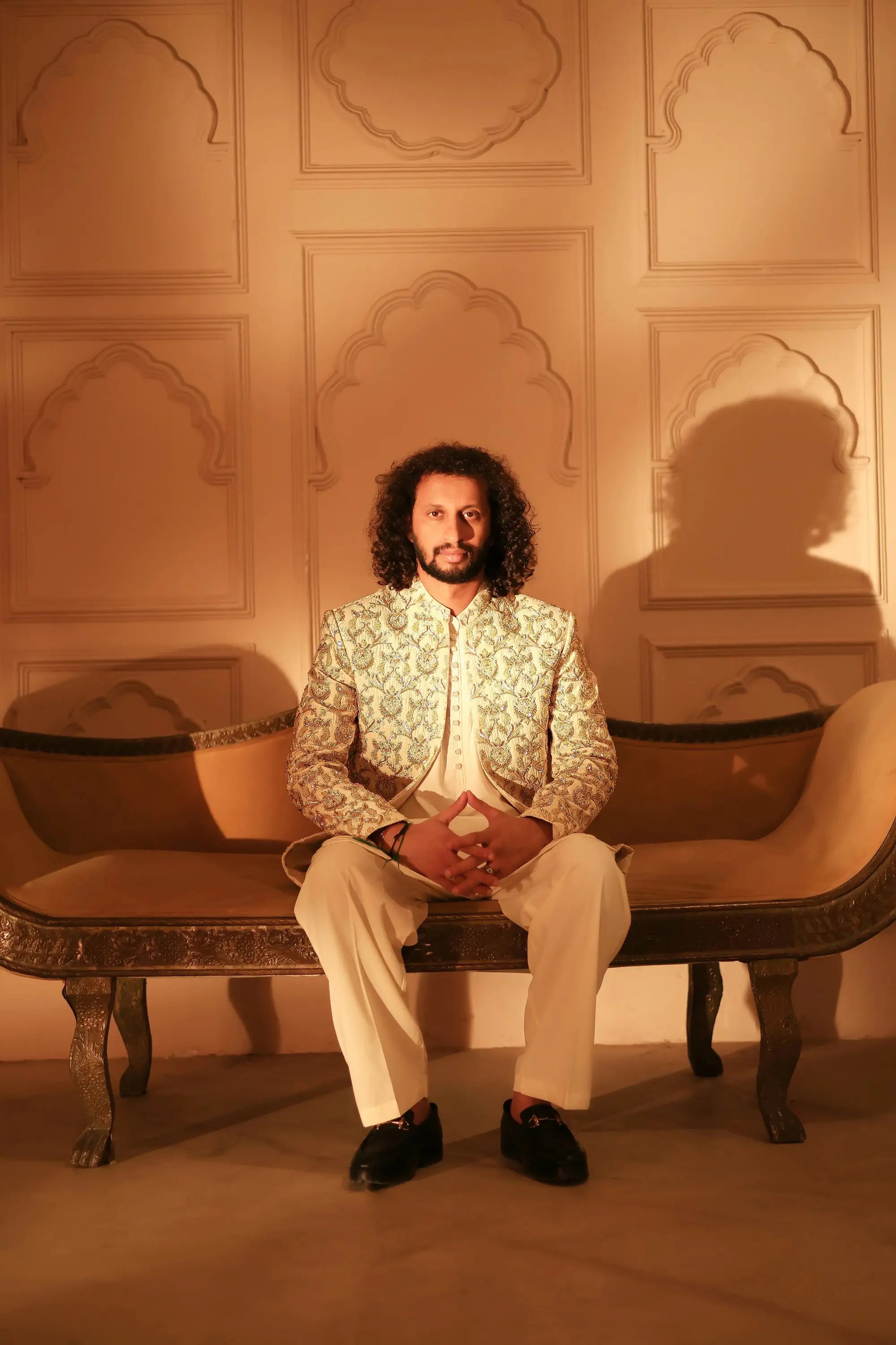 A man sits on an ornate bench wearing a cream-colored sultan jacket with intricate gold thread embroidery