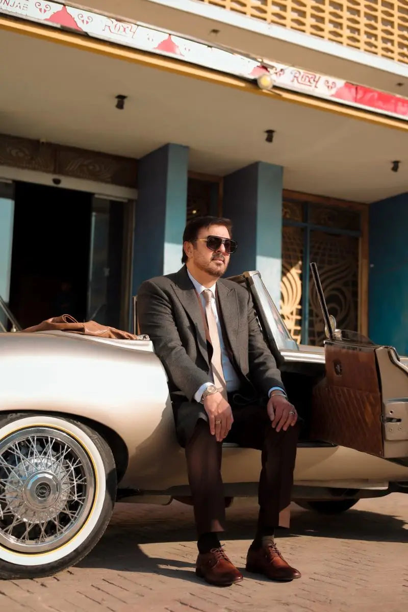 A man in a dark gray suit and brown shoes sits beside a vintage cream convertible with chrome wire wheels in waheed murad virsa-e-sitaaran edit 26
