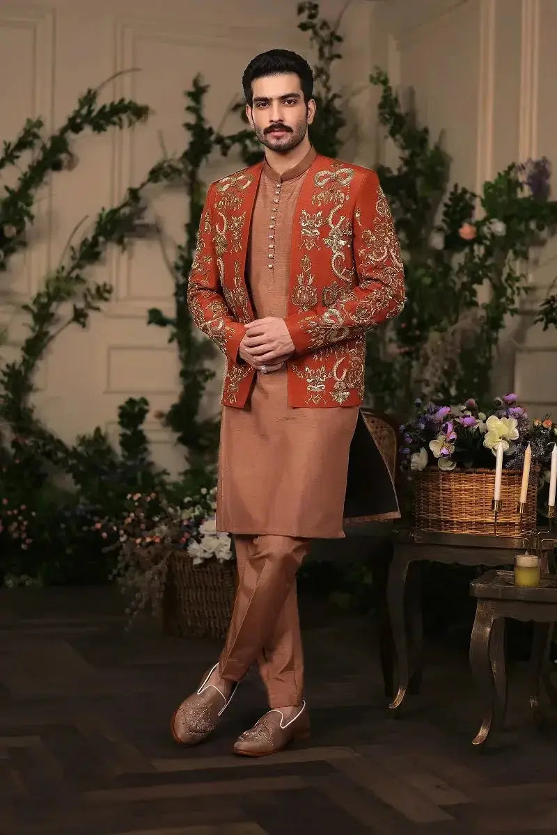 A man wearing a richly embroidered orange Wazir Khan jacket with intricate gold threadwork and matching tan trousers