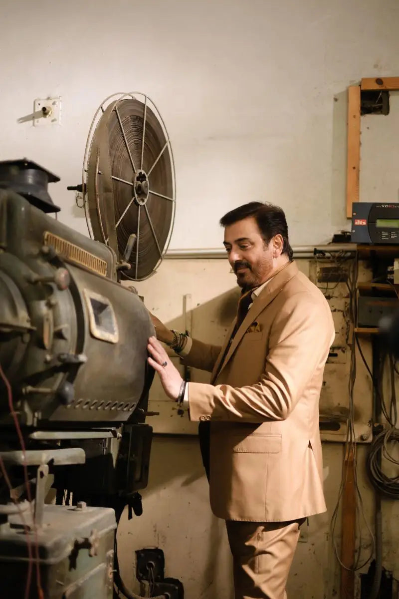 A man in a tan designer 3-piece suit for men stands beside an old film projector showcasing luxury pakistani groom wear