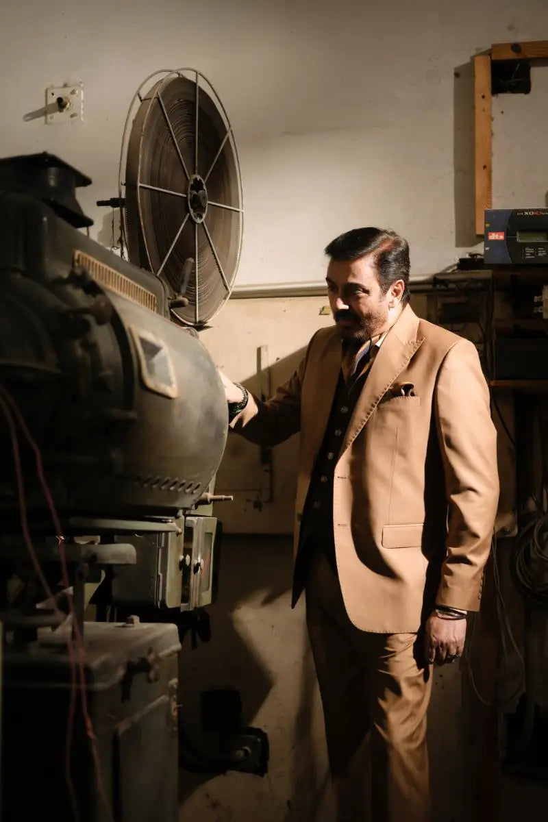 A man in a tan designer 3-piece suit for men stands beside an old film projector luxury pakistani groom wear