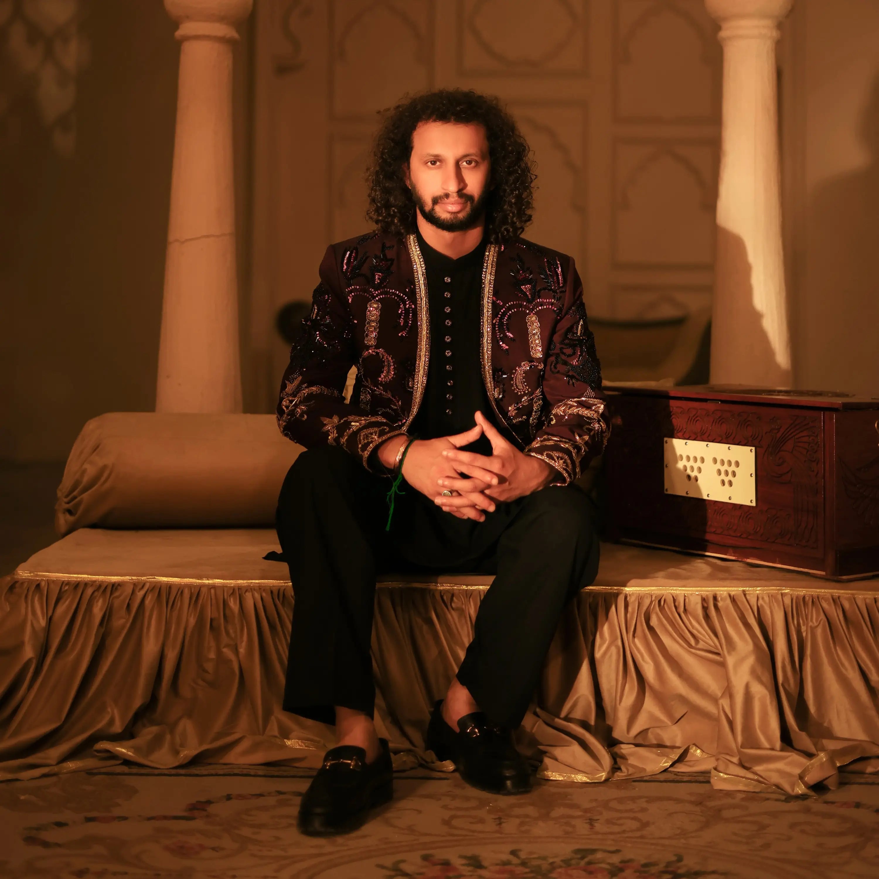 A man wears a ghazi black jacket with intricate gold embroidery in a warmly lit ornate room