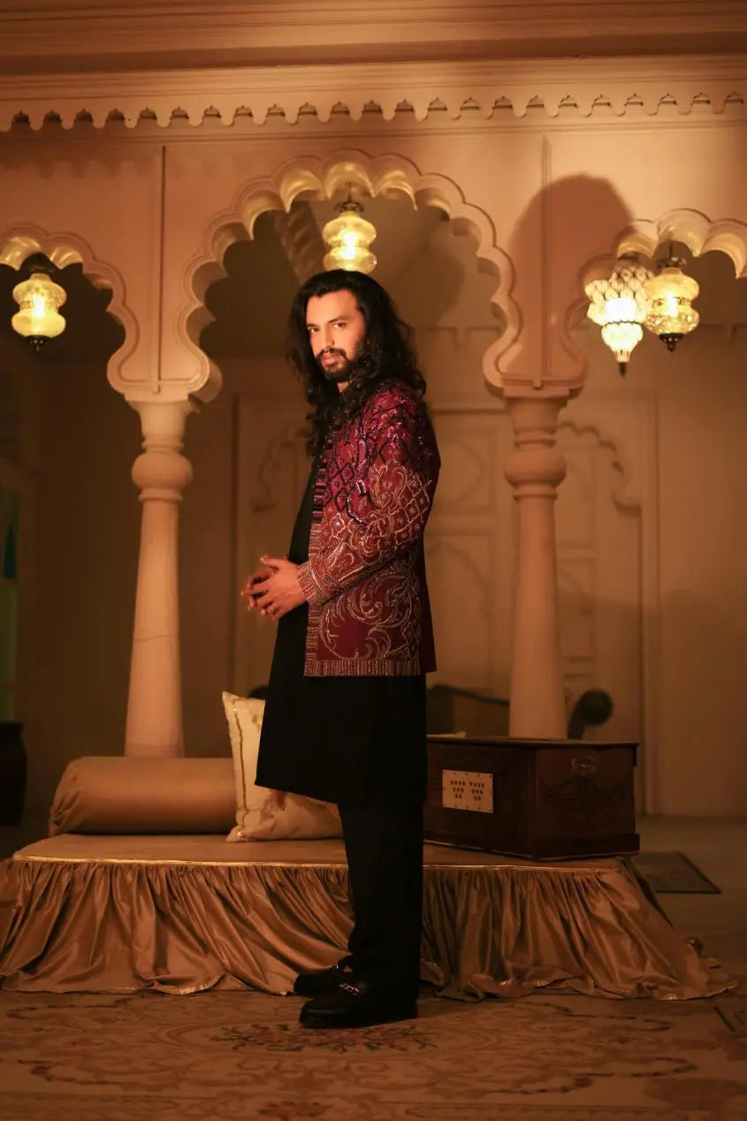 A man wearing a maroon and gold embroidered mirza jacket over black trousers showcasing intricate patterns