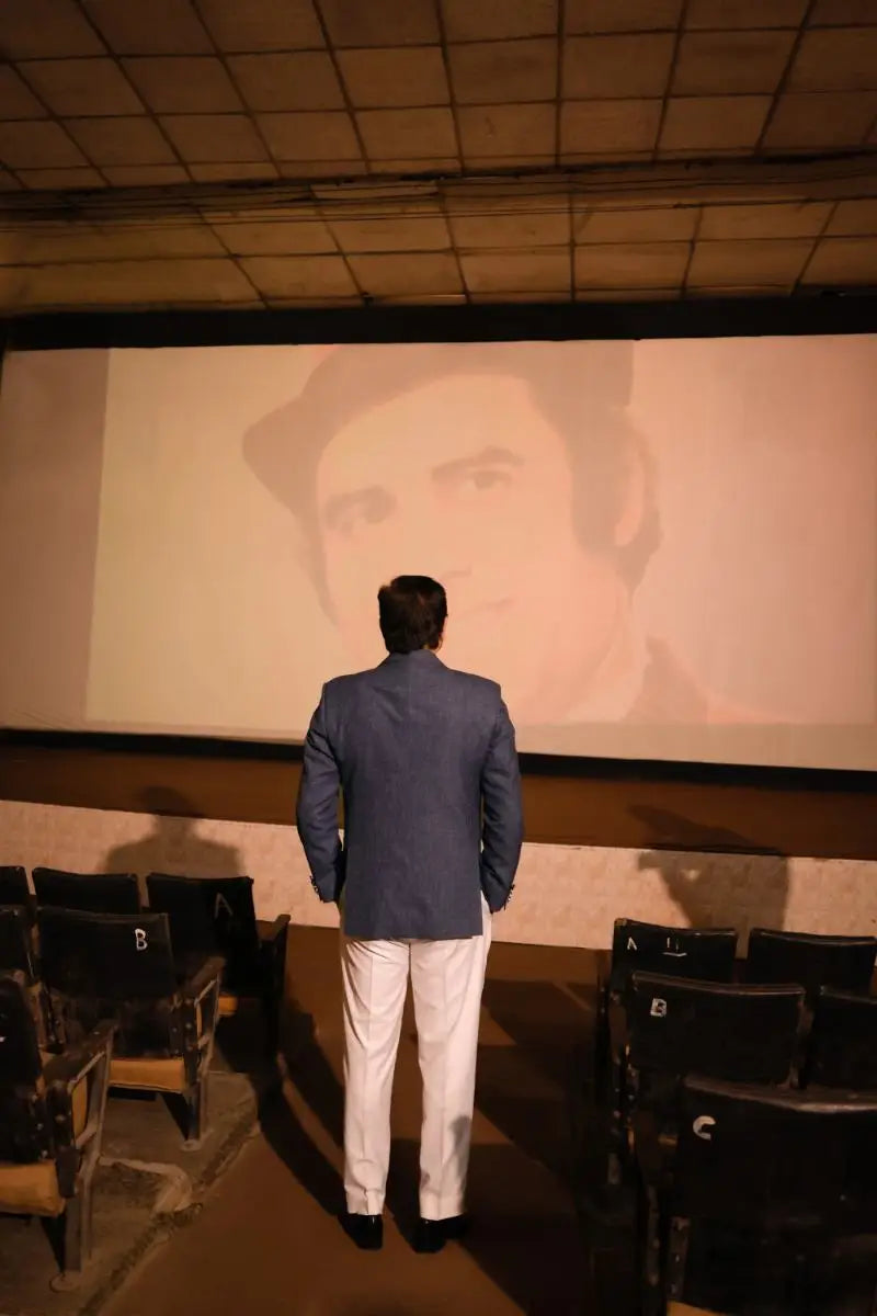 A man in a navy blue blazer and white trousers stands facing a large screen showing a face in muhammad ali virsa-e-sitaaran edit 26