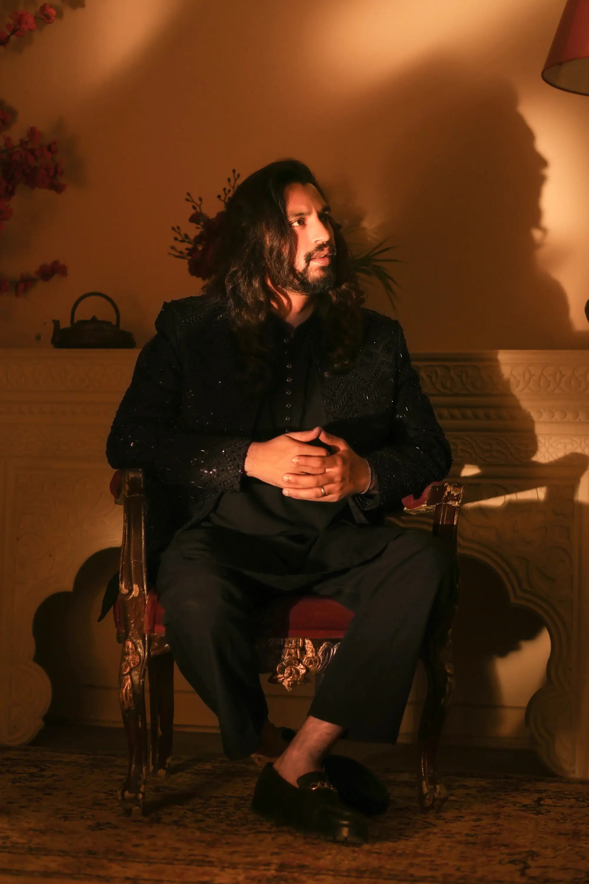 A man wearing nawab black sequined blazer and matching trousers seated in an ornate wooden chair with gold detailing