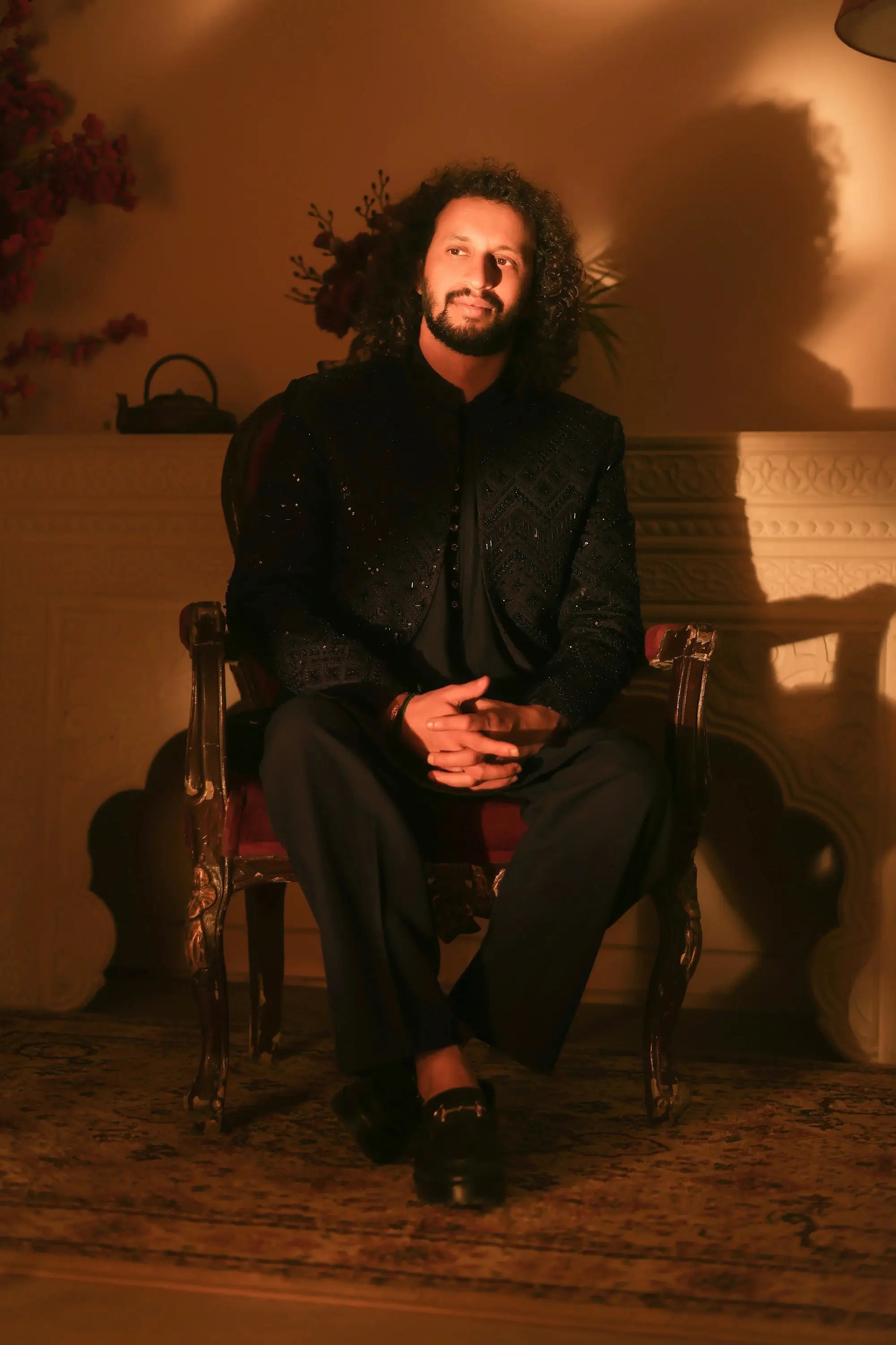 A man with curly dark hair and beard sitting in a nawab ornate carved wooden chair upholstered in deep red fabric