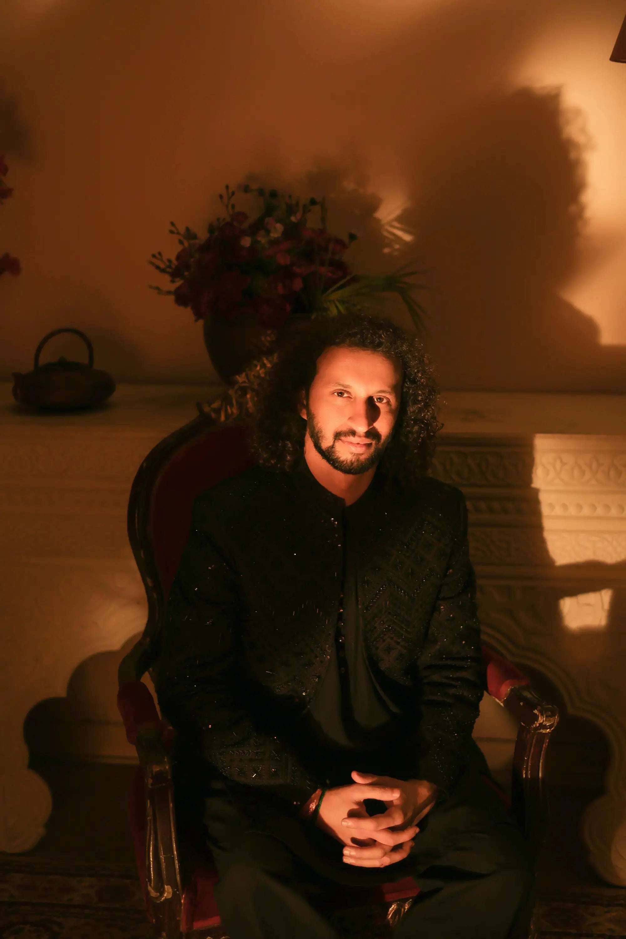 A man with long dark curly hair wearing a black formal nawab jacket with subtle sparkling embellishments sits in a red chair
