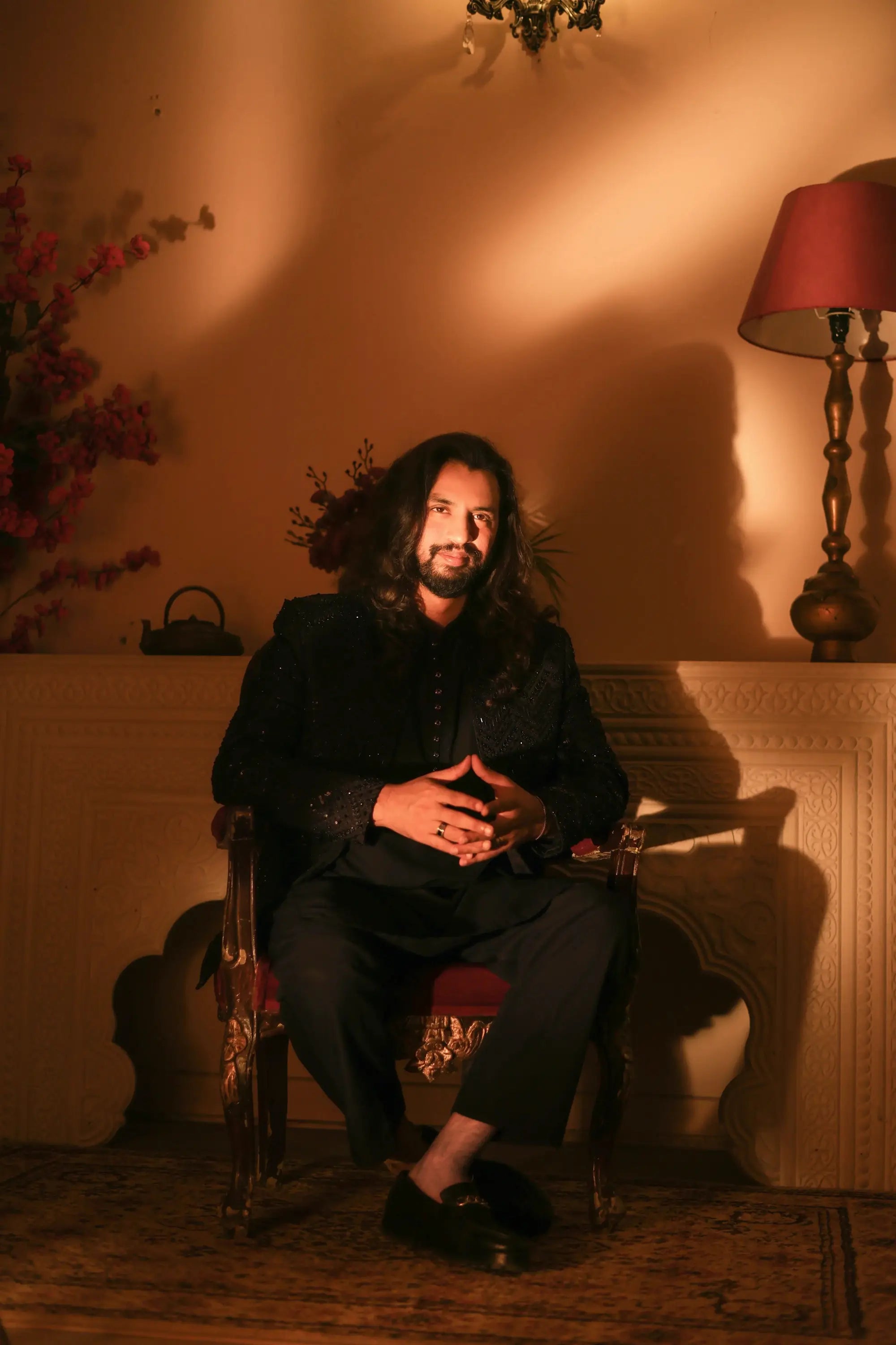 A man with long dark hair and beard sits in an ornate wooden chair wearing a black nawab formal suit with subtle embellishments