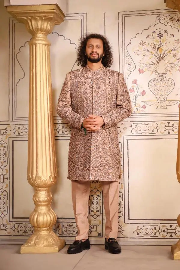 A man wearing a beige embroidered sherwani with intricate gold thread detailing and matching light brown trousers in Qalandar