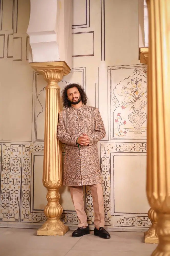 A man wearing a beige patterned traditional indian sherwani qalandar with intricate embroidery and matching light brown trousers