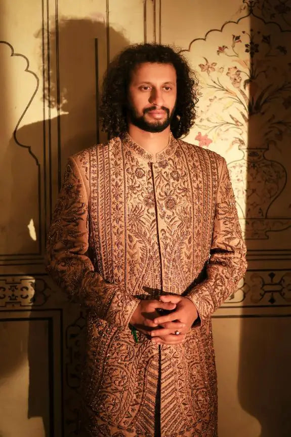 A man wearing a gold-toned qalandar sherwani with intricate embroidered paisley and floral patterns