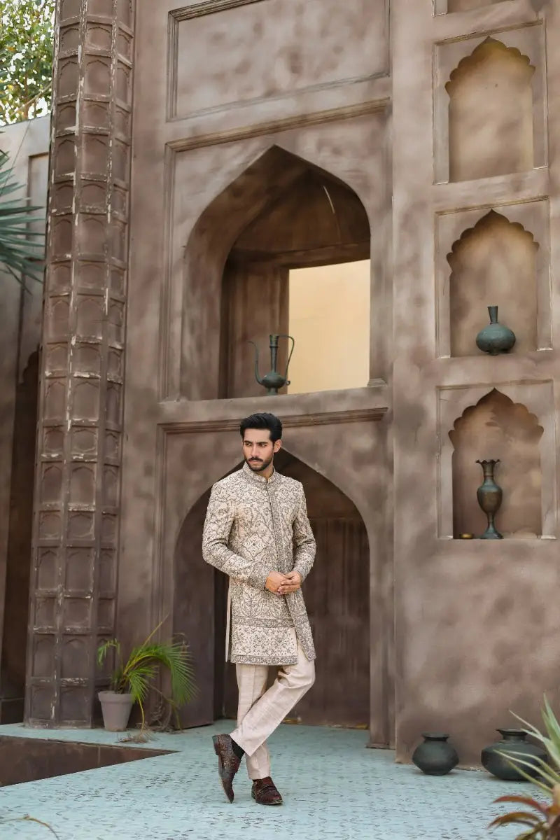 A man wearing a beige embroidered sherwani with gold thread detailing and light trousers from ahsan tariqs ishq collection qurb