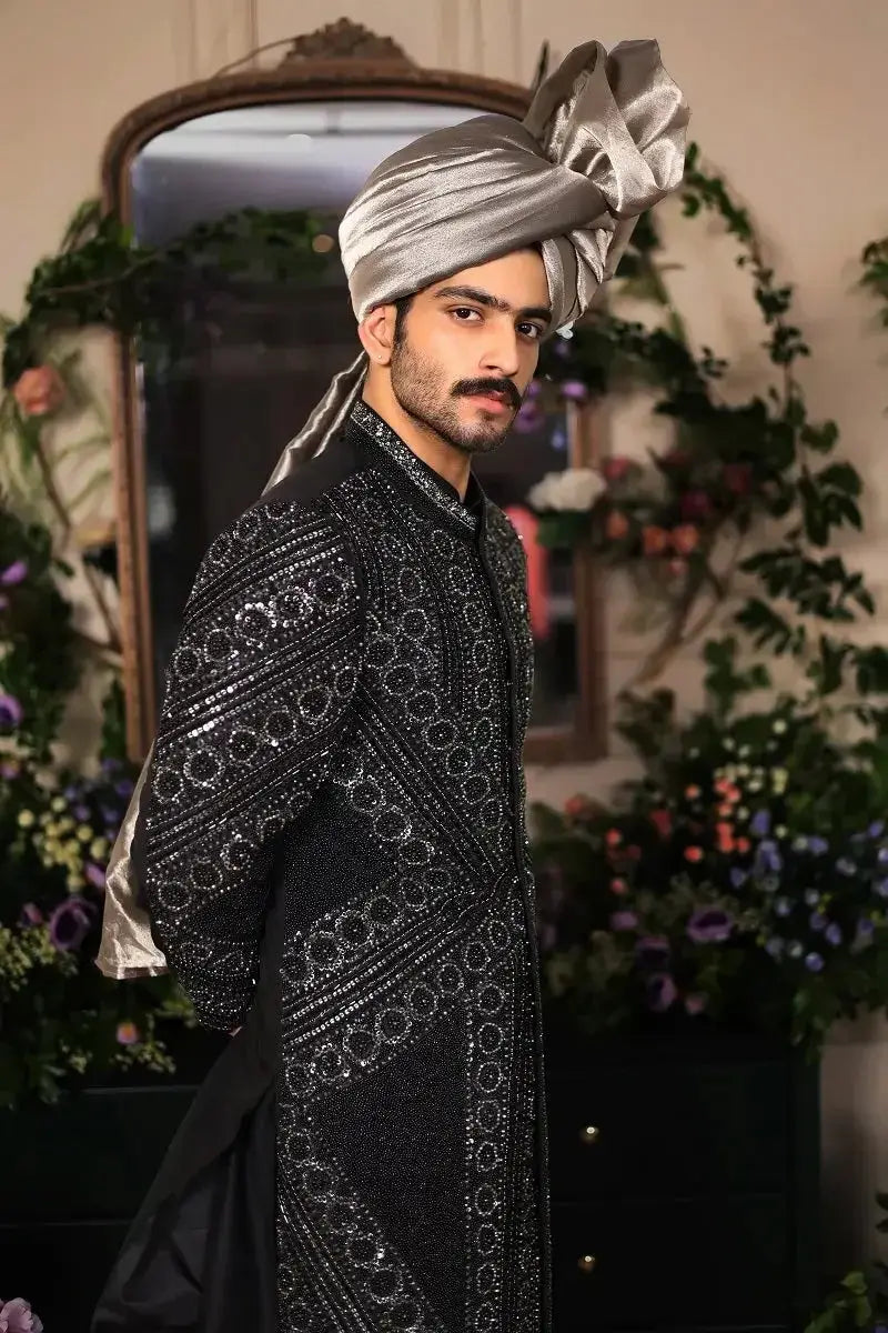 A man wears a silver satin turban with an ornate bow and black embellished sherwani in ranjeet singh royal style