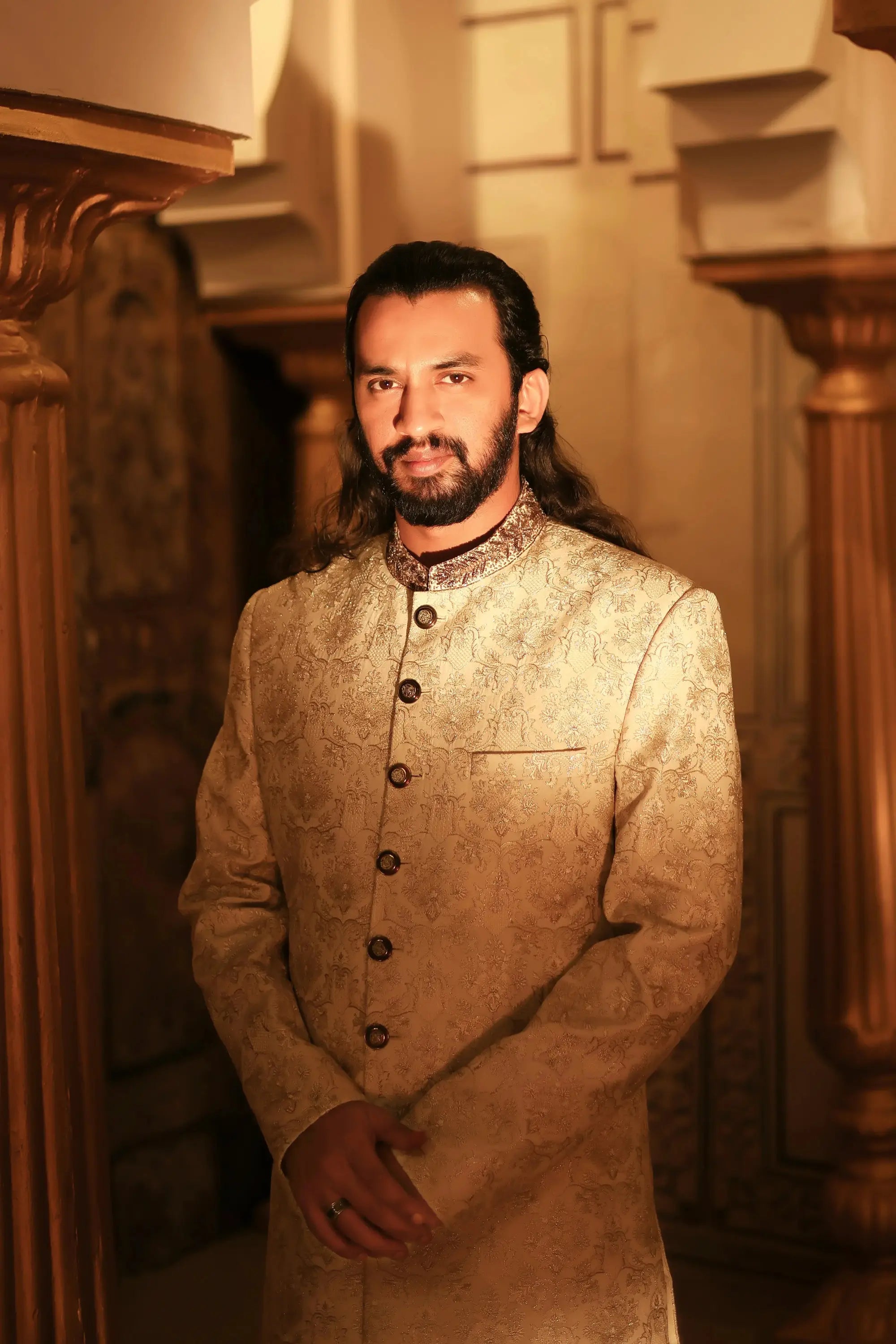 A man wearing a beige intricately patterned traditional indian sherwani with dark brown buttons and embroidery called Ranjha