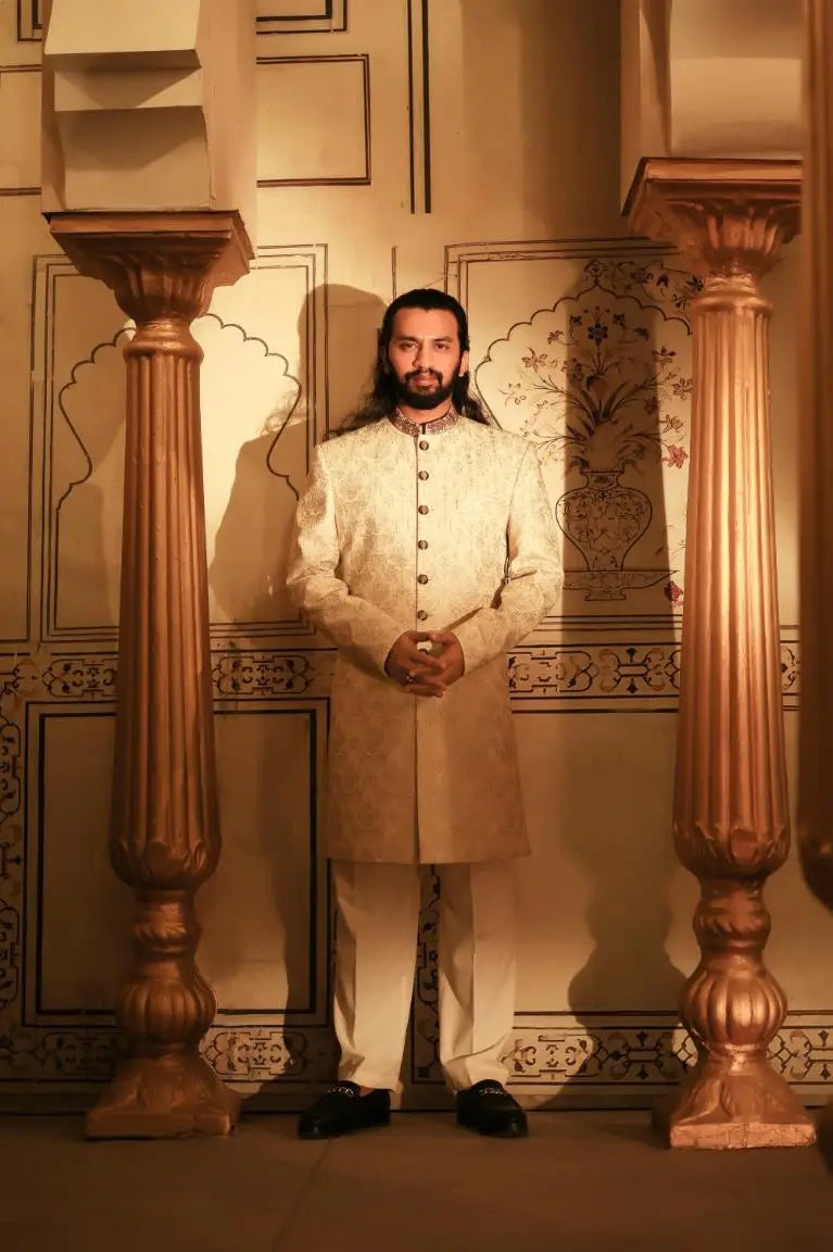 A man wearing a cream-colored embroidered sherwani with gold buttons and matching trousers from Ranjha