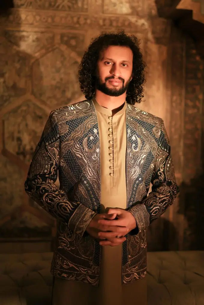 A man wearing a shayar dark blue and gold paisley jacket with intricate embroidery over a beige button-up shirt