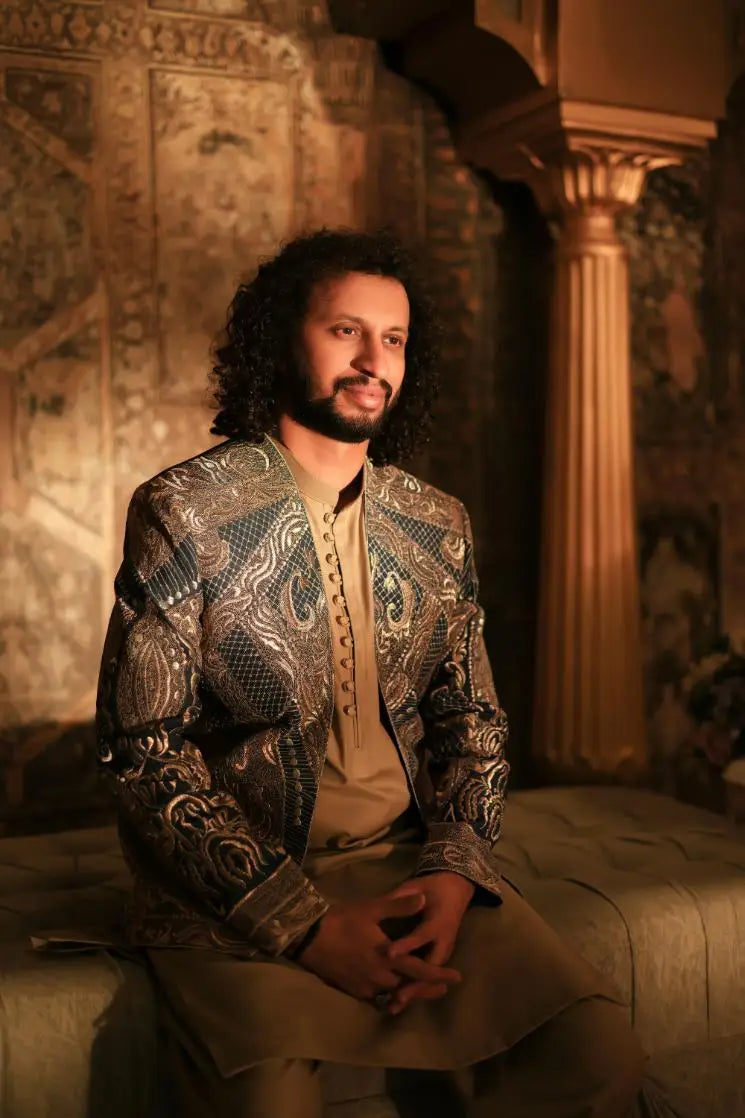 A man wears a shayar dark toned jacket with intricate gold and silver embroidery over a beige button-up shirt