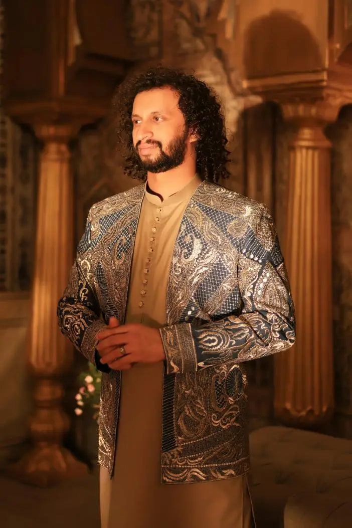 The man wears a shayar jacket with rich metallic gold and navy blue embroidery over a beige tunic