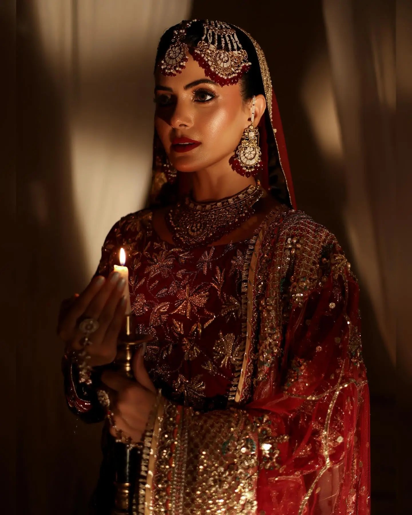 A woman in a maroon raw silk shehnai peshwas bridal outfit with antique gold vine patterns and intricate jewelry holding a candle