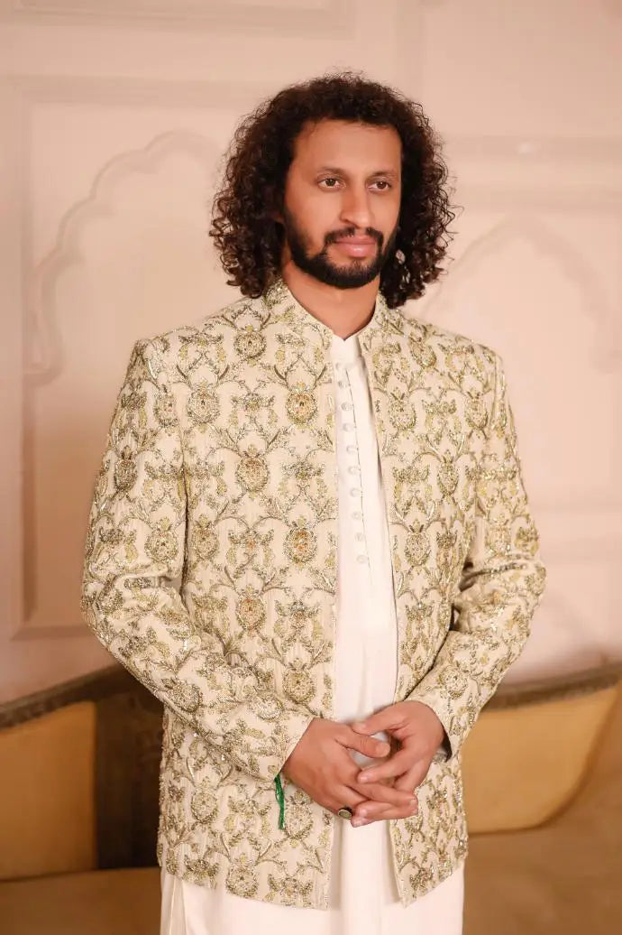 A man wearing a sultan jacket with intricate gold and green floral embroidery on cream-colored fabric