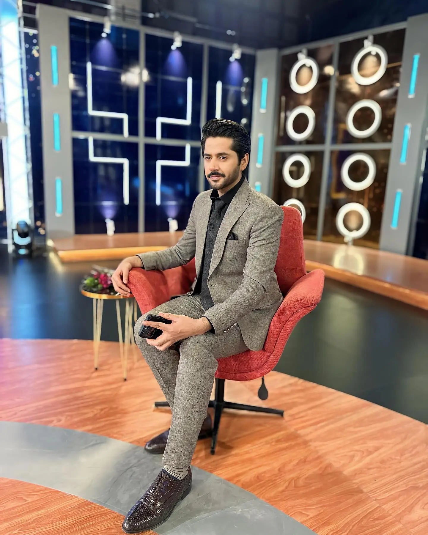 A man wears a tailored grey suit offers with textured noir fabric sitting on a red armchair by ahsan tariq