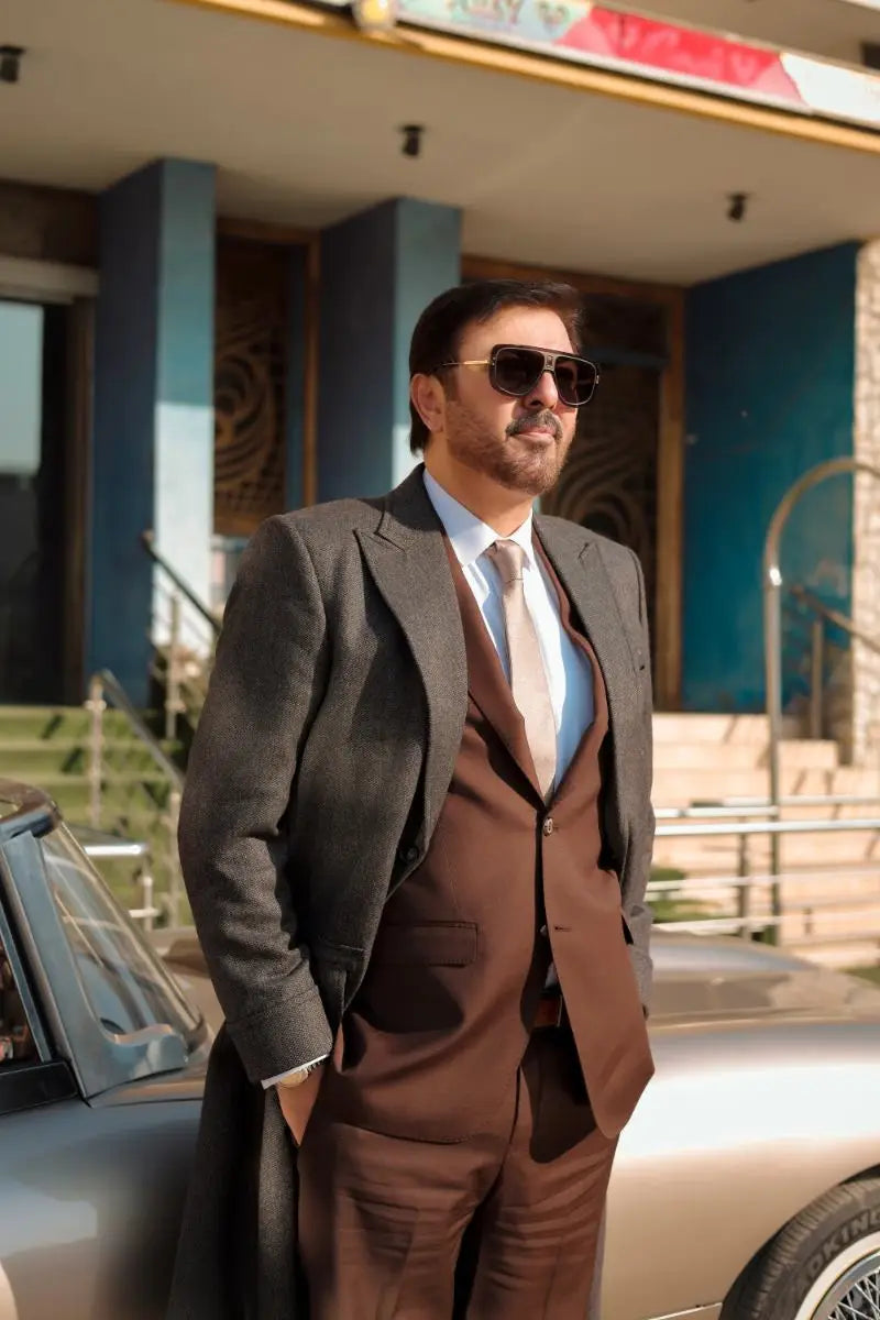 A man wearing waheed murad virsa-e-sitaaran edit 26 dark gray overcoat and brown three-piece suit with beige tie and sunglasses