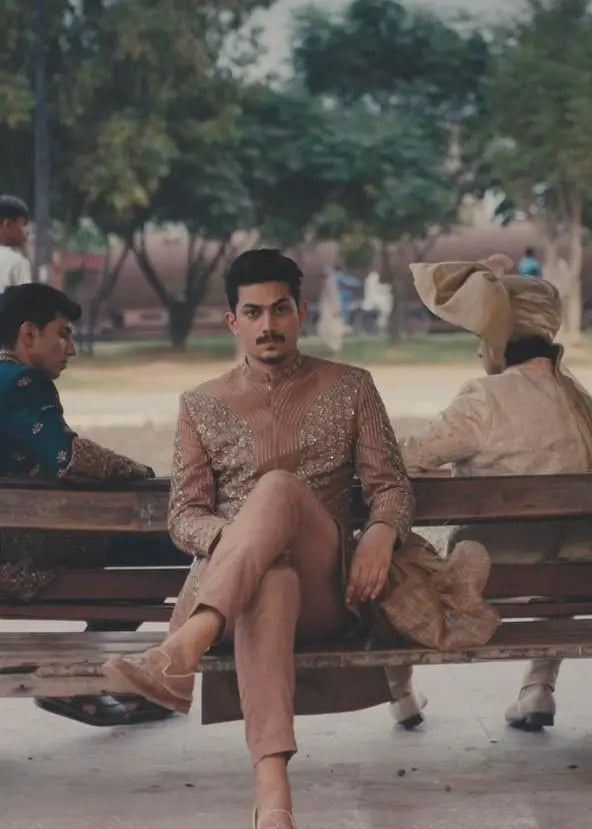 A man wearing a beige intricately embroidered zarposh traditional outfit sitting on a wooden bench